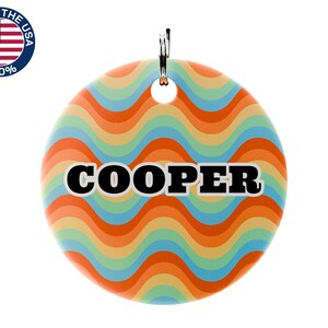 May include: A round pet tag with a retro wavy pattern in orange, yellow, blue, and green. The name "COOPER" is printed in bold black letters with a white outline. A metal ring is attached at the top. The tag is marked "Made in the USA".