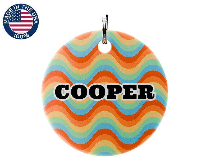 May include: A round pet tag with a retro wavy pattern in orange, yellow, blue, and green. The name "COOPER" is printed in bold black letters with a white outline. A metal ring is attached at the top. The tag is marked "Made in the USA".