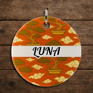 May include: A round, orange pet tag with a bonsai tree and cloud pattern. The tag has a white band with the name "LUNA" in black script. A silver ring is at the top for attachment.