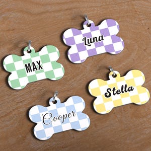 May include: Four bone-shaped pet tags with names: "Max", "Luna", "Stella", and "Cooper". Each tag has a checkered pattern in different colors: green, purple, yellow, and blue. They have a silver ring for attachment.