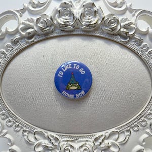 May include: A blue button with the text "I'D LIKE TO GO HOME NOW" encircling a grumpy frog wearing a party hat. The button is set against a white background within a decorative white frame.