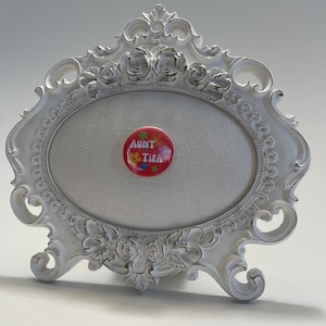 May include: A white, ornate picture frame with an oval opening, featuring floral and scroll details. A red button with the text "AUNT TIFIA" and floral accents is pinned to the center. The frame has a distressed, antique finish.