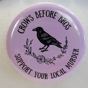 May include: A round, light purple button with black text and a black crow illustration. The text reads "CROWS BEFORE BROS" at the top and "SUPPORT YOUR LOCAL MURDER" at the bottom. The crow is centered, surrounded by floral elements.