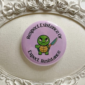 May include: A round, light purple button with the text "Respect Existence or Expect Resistance" encircling a cartoon turtle holding a knife. The turtle is green and yellow. The button is set in a white, ornate frame.