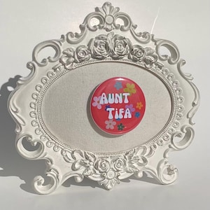 May include: A white, ornate picture frame with a canvas insert. A red button with the text "AUNT TIEA" and colorful flower graphics is centered on the canvas. The frame has floral and scroll details.