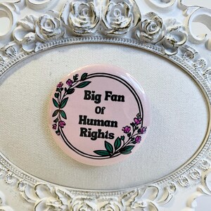 May include: A round, pink button with the text "Big Fan of Human Rights" in black, surrounded by a black border and floral accents. The button is set inside a white, ornate, oval frame.