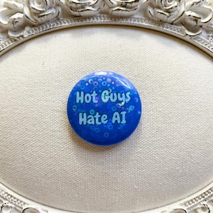 May include: A circular blue pin-back button featuring a holographic glitter finish and the text "Hot Guys Hate AI" printed in a light blue sans-serif font. The button has a smooth, reflective surface and a standard round shape.