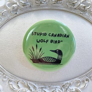 May include: A round, green button with the text "STUPID CANADIAN WOLF BIRD" in black. The button features a black and brown illustration of a loon and cattails. The button is set against a white, ornate, oval-shaped frame.