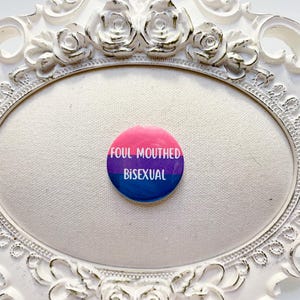 May include: A round button with a pink, purple, and blue gradient, featuring the words "FOUL MOUTHED BISEXUAL" in white. The button is set against a white oval background within a decorative white frame.