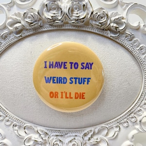 May include: A round, light yellow button with the text "I HAVE TO SAY WEIRD STUFF OR I'LL DIE" in blue and orange lettering. The button is set inside a white, ornate, oval-shaped frame with floral detailing.