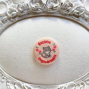 May include: A round, light pink button with the words "Become Ungovernable" in red. A gray cartoon cat is in the center, surrounded by red flowers. The button is displayed on a white oval-shaped surface within a decorative white frame.