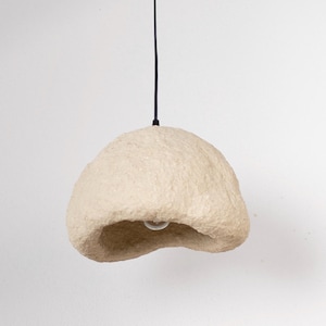 Puede incluir: Lámpara colgante beige con forma orgánica. La lámpara tiene una superficie texturizada y una bombilla visible en la parte inferior. Cuelga de un cable negro sobre un fondo blanco. La lámpara tiene una estética única y natural.