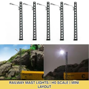 May include: Set of five black railway mast lights with a silver light fixture. The lights are designed with a lattice-style structure. The image also shows a miniature railway scene with a yellow train car and a mountain backdrop. Text at the bottom reads: RAILWAY MAST LIGHTS | H0 SCALE | MINI LAYOUT.