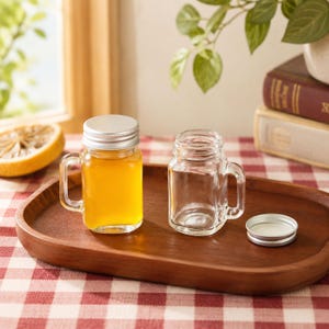 May include: Two small clear glass mason jar mugs with handles. One jar is filled with a golden yellow liquid and topped with a silver metal screw-on lid. The second jar is empty and sits next to its detached silver metal lid on a wooden tray.