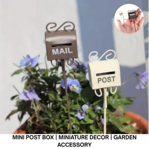 May include: Two miniature metal mailboxes, one brown with "MAIL" and the other off-white with "POST", are displayed in a flower pot with purple and blue flowers. The mailboxes have decorative scrollwork and are garden accessories.