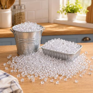 Könnte beinhalten: Ein Eimer aus verzinktem Metall und eine rechteckige Kastenform aus Metall, gefüllt mit kleinen, klaren, facettierten Acrylperlen. Die Perlen sind auch auf der Holzoberfläche vor den Behältern verstreut und bilden einen Haufen reflektierender, diamantartiger Teile.