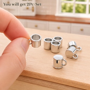 May include: A set of miniature silver-colored metal mugs with a smooth, reflective finish and a small handle on the side. The text "You will get 2Pc/Set" is visible at the top of the image. These tiny cups are designed for small-scale displays.
