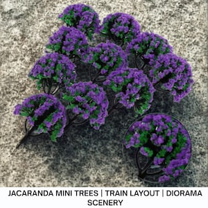 May include: Miniature Jacaranda trees with purple and green foliage, ideal for train layouts and dioramas. The trees have dark brown trunks and branches. The image includes a close-up of the tree's foliage. Text on the image reads: JACARANDA MINI TREES | TRAIN LAYOUT | DIORAMA SCENERY.
