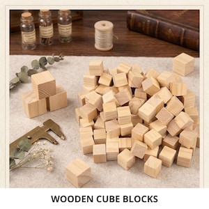 May include: A large collection of small, unfinished light-coloured wooden cube blocks arranged in a pile, with several individual cubes placed nearby, a metal calliper tool, and sprigs of dried greenery on a neutral fabric surface.