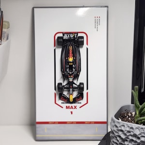 Può includere: Modello di auto da corsa Red Bull Racing Max 1, con il testo "BYBIT" e "PIT LANE". L'auto è nera, rossa e gialla, su sfondo bianco. Il modello è esposto su una superficie bianca.