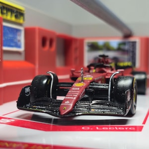 Peut inclure: Une maquette rouge et noire de voiture de course de Formule 1 avec des logos de sponsors, garée dans un décor de garage. La voiture a de grands pneus noirs et un aileron avant proéminent. L'arrière-plan comprend des murs rouges et une enseigne Ferrari.