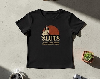 May include: Black ribbed crew neck t-shirt with the word "SLUTS" in a retro font, a graphic of a woman, and the text "Sexy Ladies Under Tremendous Stress." Sunglasses, a camera, and a small plant are also visible.