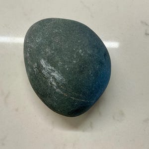 Peut inclure: Une pierre de rivière arrondie et lisse de couleur gris foncé avec une texture mate. La pierre présente une fine veine minérale blanche subtile qui traverse sa surface en diagonale, créant un contraste naturel avec le gris ardoise.