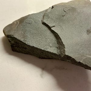 Könnte beinhalten: Ein Stück graues Schiefergestein mit rauer, strukturierter Oberfläche. Der Stein hat ein geschichtetes Aussehen und eine gezackte Kante, mit einem sichtbaren Riss, der durch die Mitte verläuft. Der Stein ist vor einem weißen Hintergrund platziert.