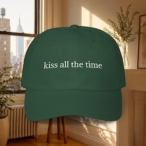 May include: A dark green baseball cap featuring the white embroidered text "kiss all the time" centered on the front panel. The cap has a structured crown, a curved brim, and a button at the top, presenting a clean and simple design.