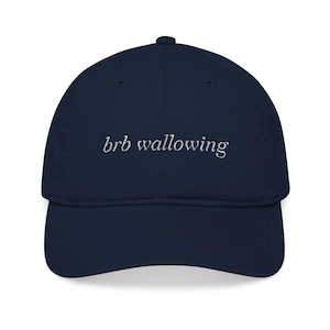 May include: A navy blue baseball cap featuring the embroidered white text "brb wallowing" centered on the front panel. The cap has a structured crown, a curved brim, and a button at the top, showcasing a clean and simple design.