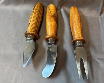 May include: Three cheese knives with wooden handles and stainless steel blades. The handles are a light brown color with a wood grain pattern. One knife has a pointed blade, one has a curved blade, and one has a two-pronged fork.