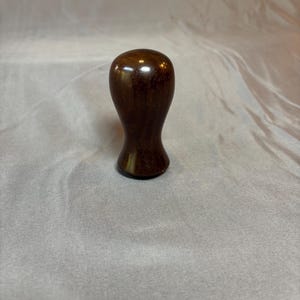 Könnte beinhalten: Kleines, dunkelbraunes Holzobjekt mit abgerundeter Oberseite und schmalerer Basis. Das Holz hat eine polierte, glänzende Oberfläche, die das Licht reflektiert. Das Objekt scheint ein Griff oder Knopf zu sein, möglicherweise für eine Schublade oder einen Schrank.