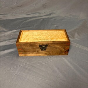 May include: A rectangular wooden box with a light-colored top and a darker, patterned body. The box features a decorative metal clasp and small orange accents. The wood grain is visible, adding to the handcrafted aesthetic.