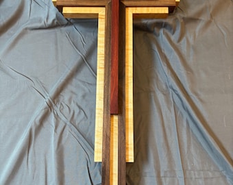 May include: A wooden cross with a layered design. The cross features a central red-brown cross shape, surrounded by layers of light and dark wood. The design is a mix of vertical and horizontal elements, creating a dimensional effect.