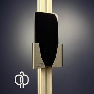 Peut inclure: Un support de téléphone argenté monté sur un poteau vertical. Le support a une ouverture en forme de V pour fixer un smartphone noir. Le support porte les initiales "LM" sur le devant. Le fond est un dégradé de bleu foncé et de gris.