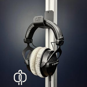 May include: Black and gray over-ear headphones hanging on a black metal wall mount. The headphones have gray ear pads and the text "DT 770 PRO" is visible. The wall mount is attached to a silver metal pole.