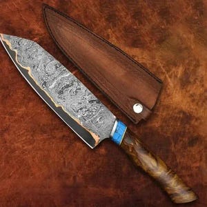 May include: A Damascus steel chef's knife with a wooden handle and a leather sheath. The blade has a unique pattern, with a gold accent along the edge. The handle features blue and wood tones.