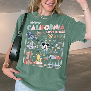 Op de afbeelding: Groen t-shirt met een afbeelding van Disney California Adventure. Het shirt heeft Mickey Mouse en verschillende parkattracties. De persoon draagt een lichtblauwe spijkerbroek en witte sneakers. De persoon draagt ook een zwarte zonnebril en een zwarte handtas.