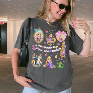 May include: A charcoal gray short-sleeve t-shirt featuring a colorful graphic print of Rapunzel, a horse, and a chameleon. The text "AT LAST I SEE THE LIGHT" is printed in the center, surrounded by various illustrations of the character in frames.