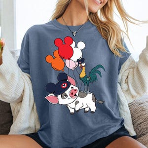 Op de afbeelding: Blauw t-shirt met een cartoonvarken met een hoed, een haan en Mickey Mouse-vormige ballonnen in rood, oranje en wit. Het varken heeft zwart-witte vlekken. De haan is groen en geel. De ballonnen worden door de haan vastgehouden.