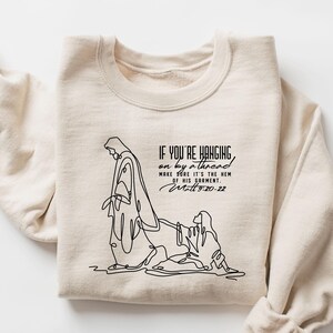 May include: Cream-colored sweatshirt featuring a black line drawing of a religious figure and text. The text reads "IF YOU'RE HANGING on by a thread MAKE SURE IT'S THE HEM OF HIS GARMENT Matthew 9:20-22."