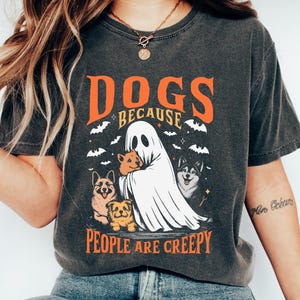 Może przedstawiać: Ciemnoszara koszulka z napisem "DOGS BECAUSE PEOPLE ARE CREEPY" w kolorze pomarańczowym i białym. Grafika przedstawia ducha trzymającego psy, z nietoperzami w tle. Koszulka ma swobodny, vintage'owy styl.