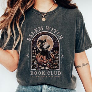 May include: Dark gray t-shirt with a Salem Witch Book Club graphic. The design features a witch reading on a pumpkin, with a full moon backdrop. The text "ALL WITCHES WELCOME" is also included.