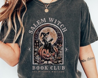 May include: Dark gray t-shirt with a Salem Witch Book Club graphic. The design features a witch reading on a pumpkin, with a full moon backdrop. The text "ALL WITCHES WELCOME" is also included.