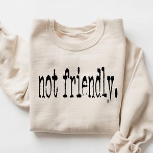 Könnte beinhalten: Cremefarbenes Sweatshirt mit dem schwarzen Aufdruck „not friendly.“ in einer stilisierten Schriftart. Die Ärmel sind hochgekrempelt, und das Sweatshirt ist gefaltet. Der Gesamteindruck ist lässig und bequem.