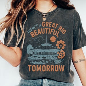 May include: Dark gray t-shirt with the text "There's a Great Big Beautiful Tomorrow" in orange. The graphic includes a building, sun, and gears. The shirt is paired with blue jeans and white sneakers.