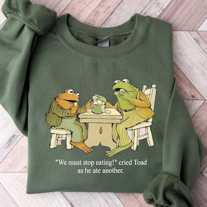 May include: Green sweatshirt featuring a cartoon illustration of two frogs at a table. The frogs are dressed in clothing and the text "We must stop eating!" is printed below the illustration. The sweatshirt has long sleeves.