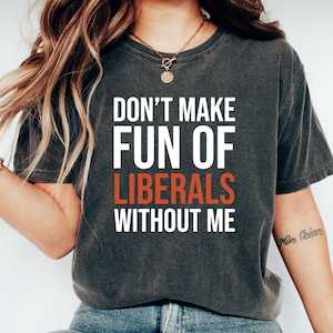 May include: Dark gray t-shirt with the text "DON'T MAKE FUN OF LIBERALS WITHOUT ME" in white and orange. The shirt is paired with blue jeans and white slip-on shoes. The overall style is casual and relaxed.