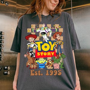 May include: Dark gray t-shirt featuring a colorful graphic of Toy Story characters, including Woody, Buzz Lightyear, and Jessie. The words "Toy Story" are in a yellow and red font, with "Est. 1995" below. The shirt has short sleeves and a relaxed fit.