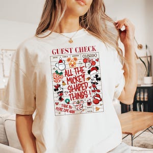 Pode incluir: Uma t-shirt de cor creme com um gráfico vermelho e preto de uma conta de restaurante. O texto diz GUEST CHECK, ALL THE MICKEY SHAPED THINGS e THANK YOU PLEASE COME AGAIN, rodeado por vários ícones do Mickey Mouse com tema natalício.